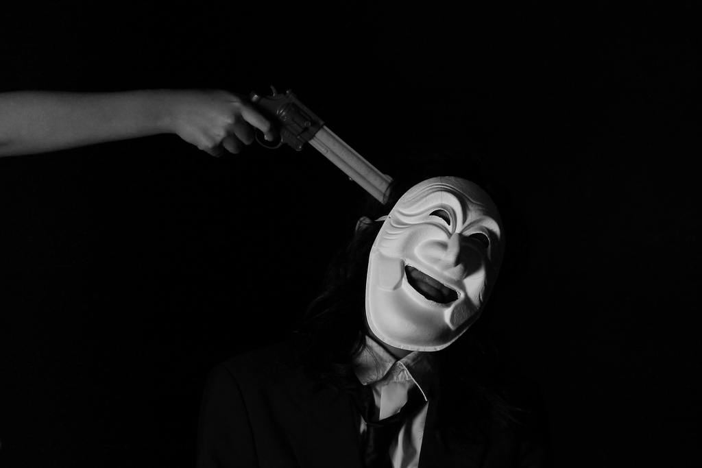 Anonymous hand pointing a gun at the man with a white smiling mask