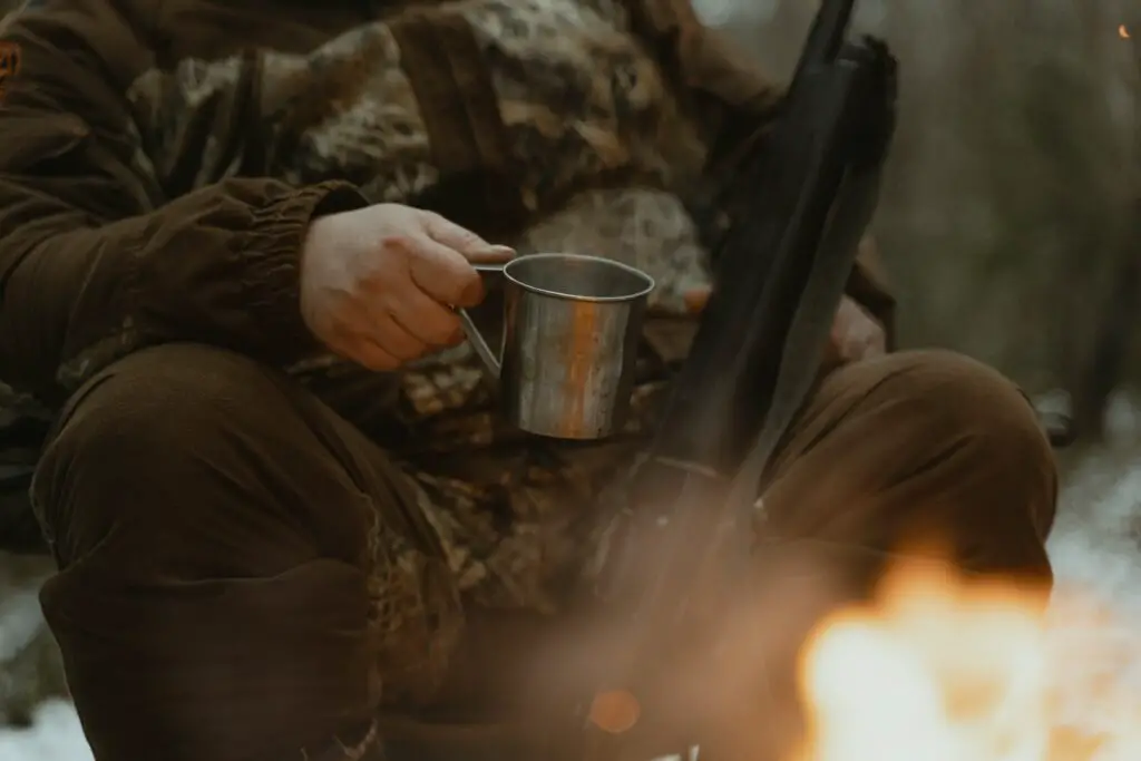 man sitting in a camp fire holding a cup and gun