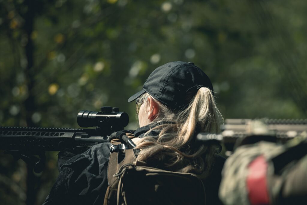 A woman waring a cap and jacket sniping through rifle scope