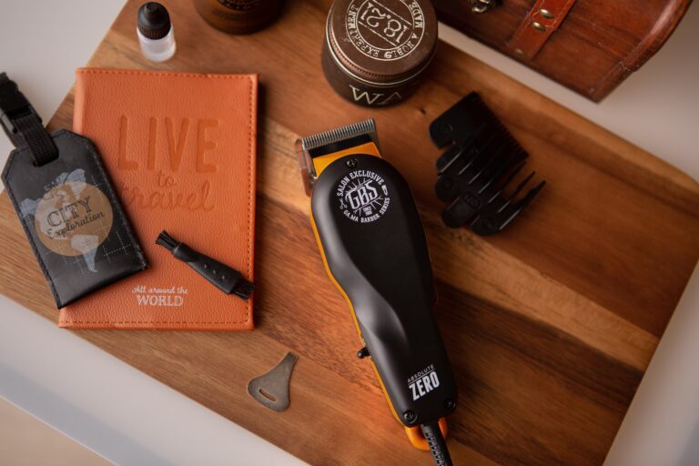 A black and orange hair clipper near an orang passport holder, black luggage tag, hair clipper oil, and pomade on top of a brown wooden board
