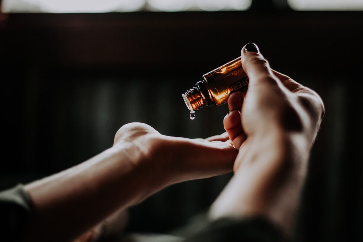 A person wearing a green long sleeves top is holding a brown bottle with oil in a dimly lit room