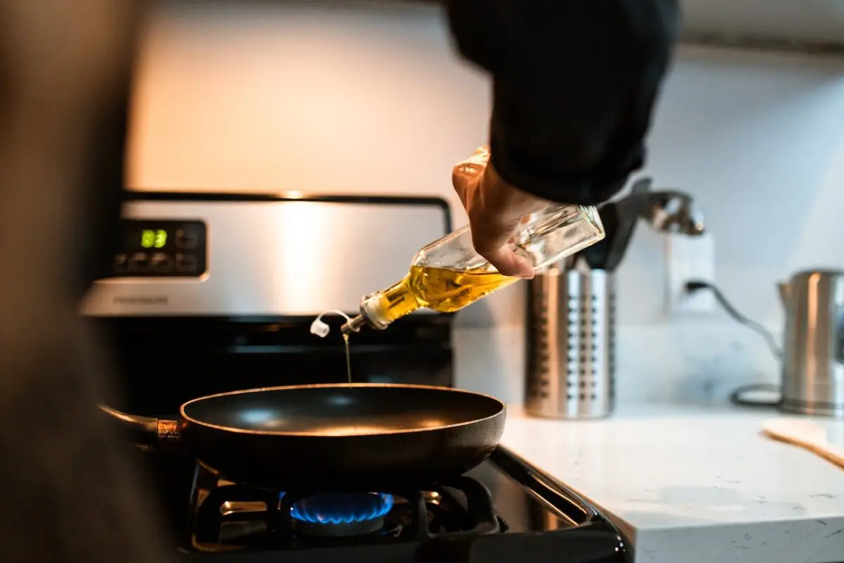 A person wearing a black long-sleeve shirt is pouring oil from a glass oil dispenser into a black frying pan