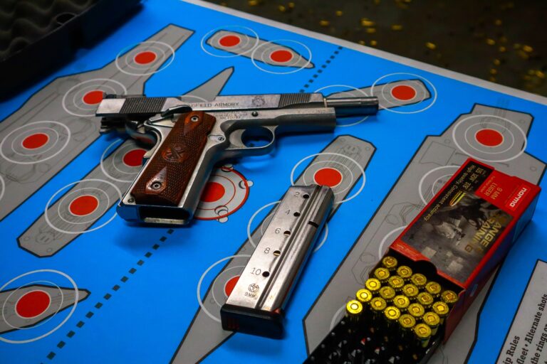 Silver and brown colored hand pistol beside a silver magazine and gold bullets on top of a blue and gray target board