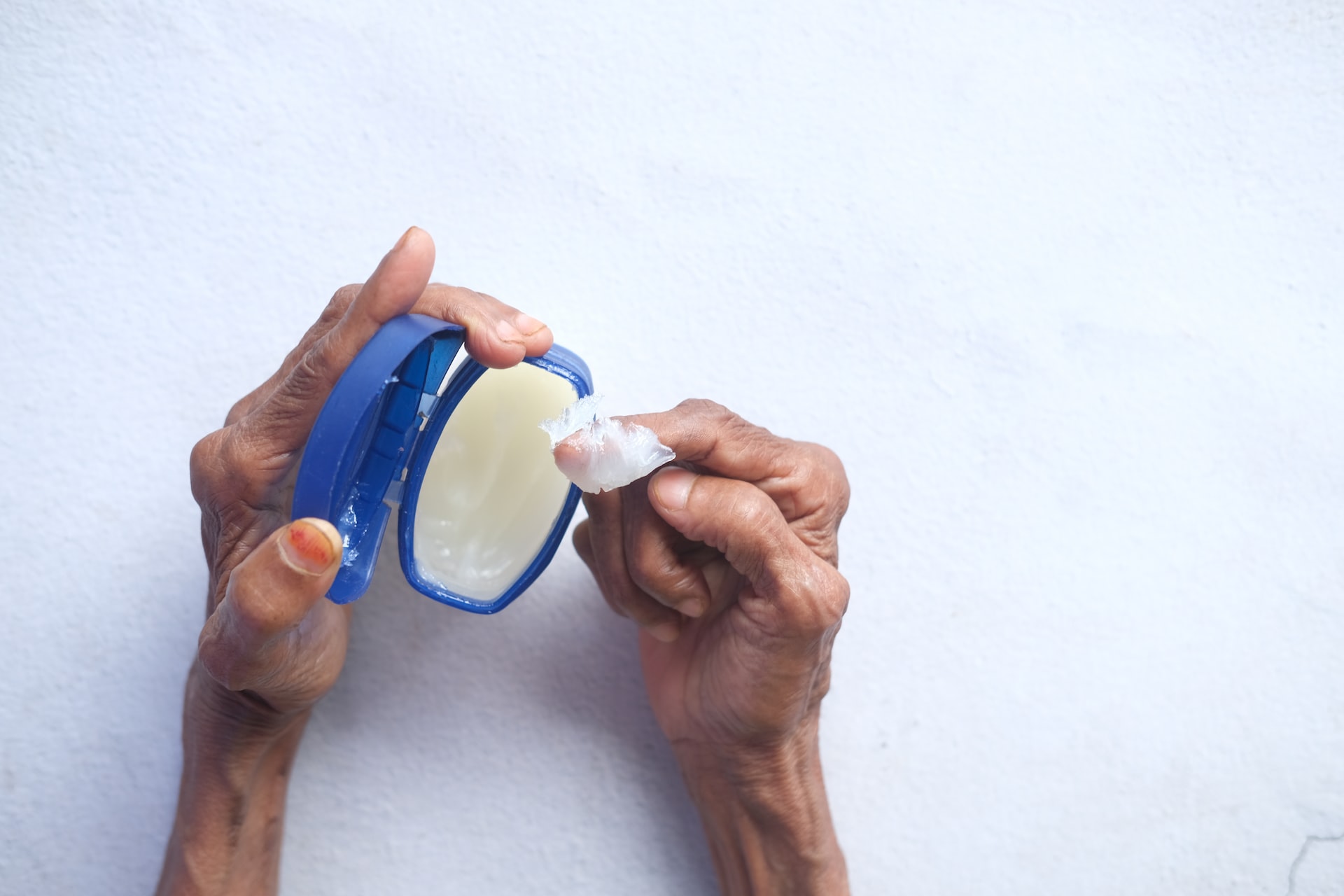 A person getting vaseline using one finger while holding a blue container near a white surface